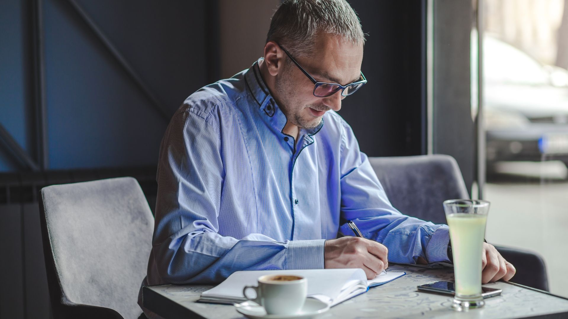 CEO escribiendo en una libreta mientras reflexiona sobre su estrategia de marca personal, en un café, con una bebida y su móvil sobre la mesa — imagen vinculada al artículo de Gabriel Patrizzi sobre por qué el CEO debe liderar su posicionamiento con independencia del equipo de comunicación corporativa